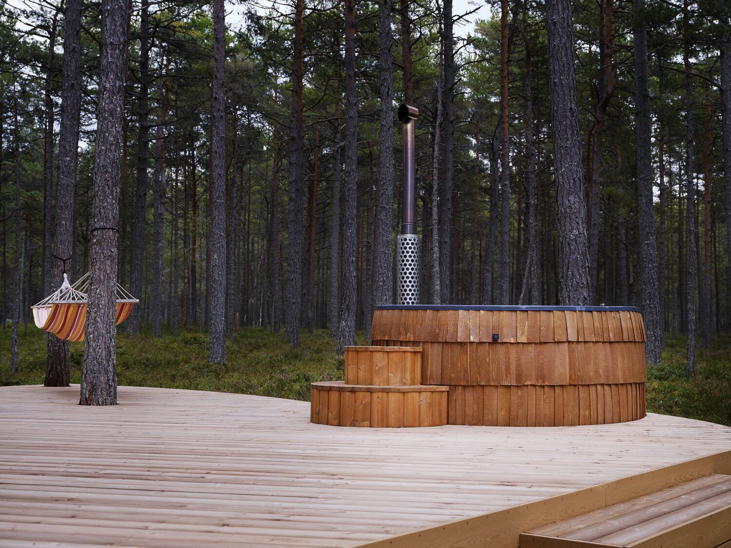 A Chestnut Iglucraft Hot Tub on a wooden deck in a pine forest at soft daylight, matching wooden steps leading up to the tub, a striped hammock strung between two trees nearby