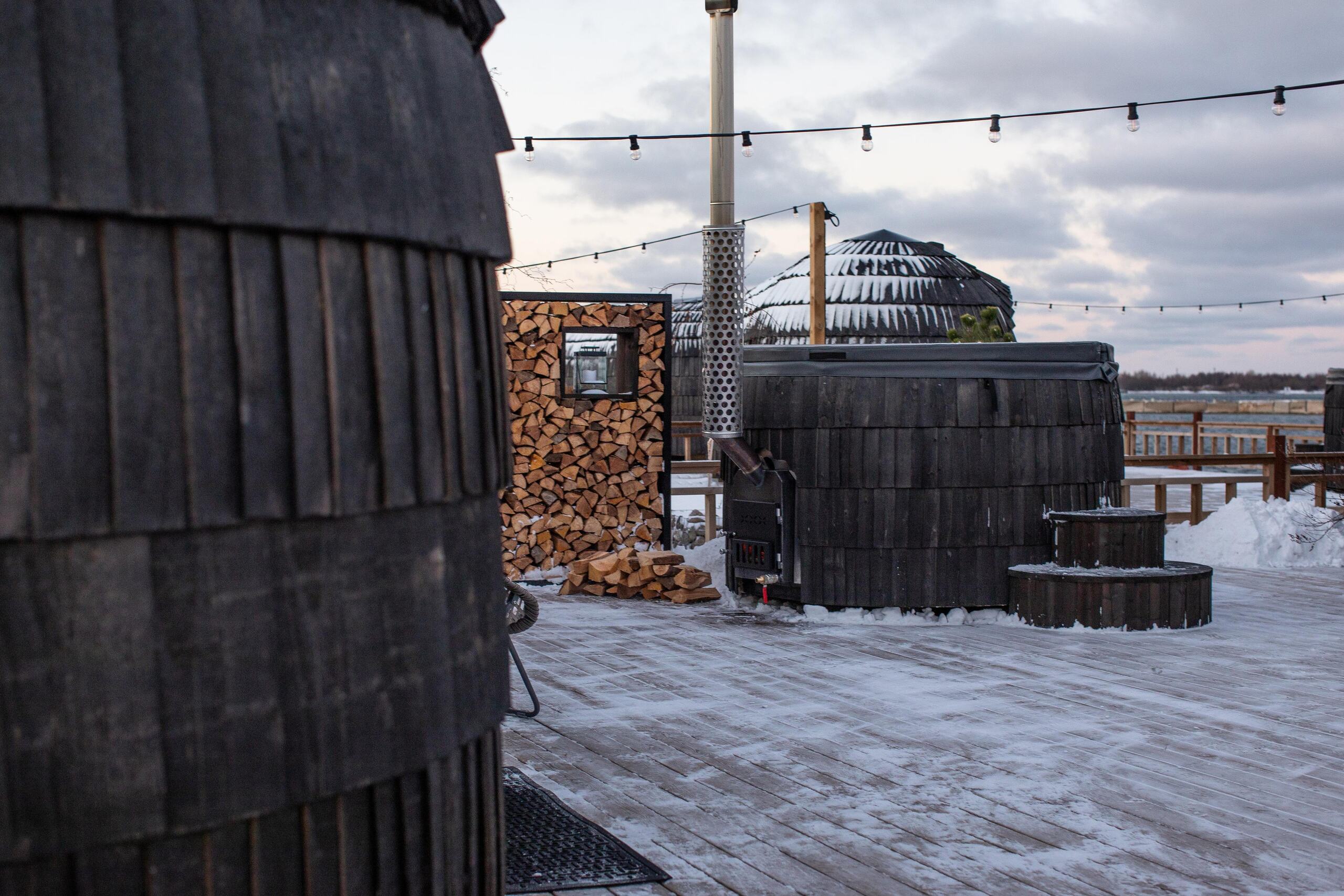 An Ebony Iglucraft Hot Tub on a snowy waterfront deck at dusk — string lights overhead, a tall stack of split firewood beside it, the wood stove door visible at its base, an Iglucraft sauna in the background under fresh snow