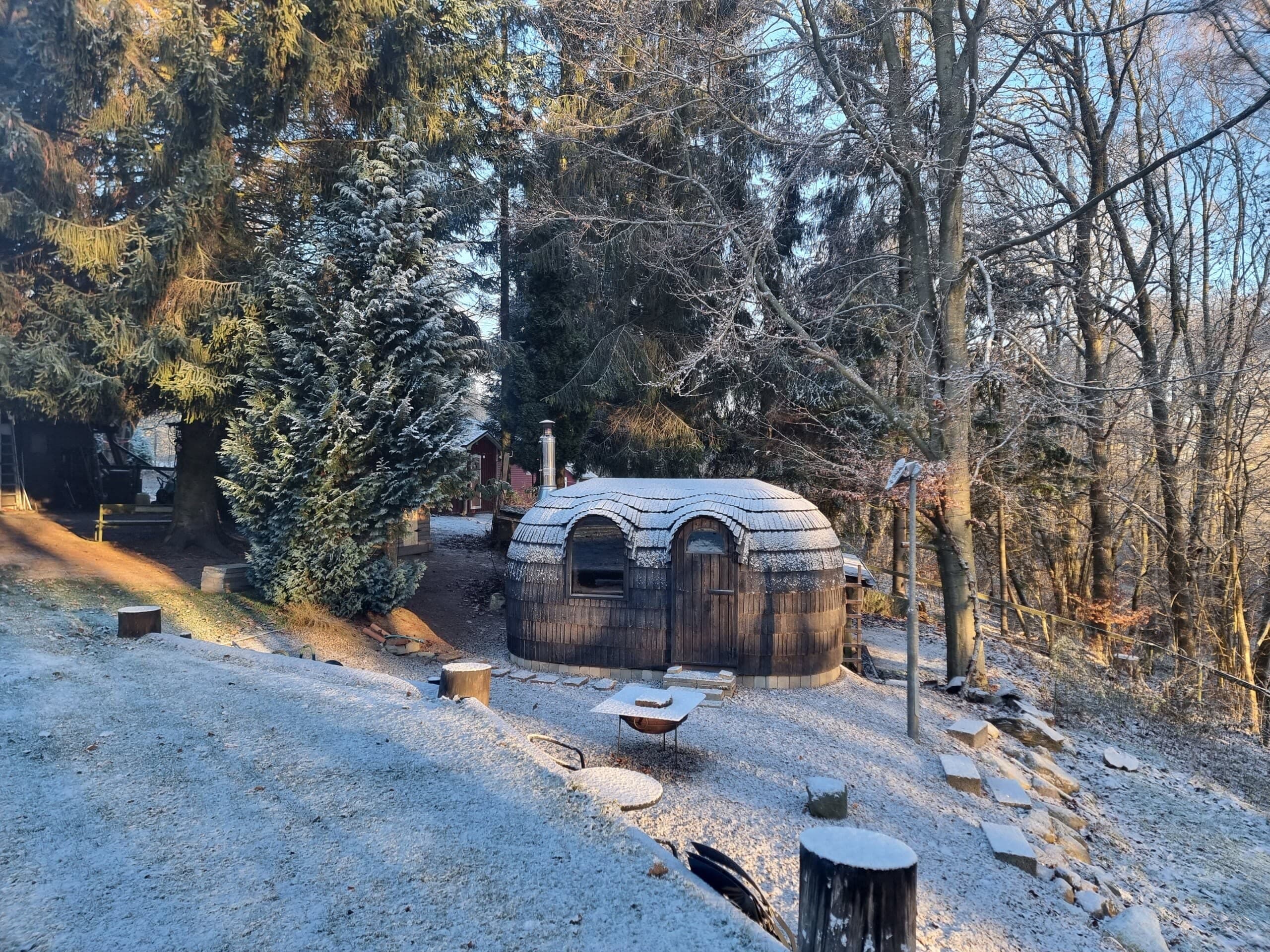 An Ebony Double Iglusauna on a frost-covered hillside in winter morning sun, evergreen trees behind, a small fire pit and red shed in the distance