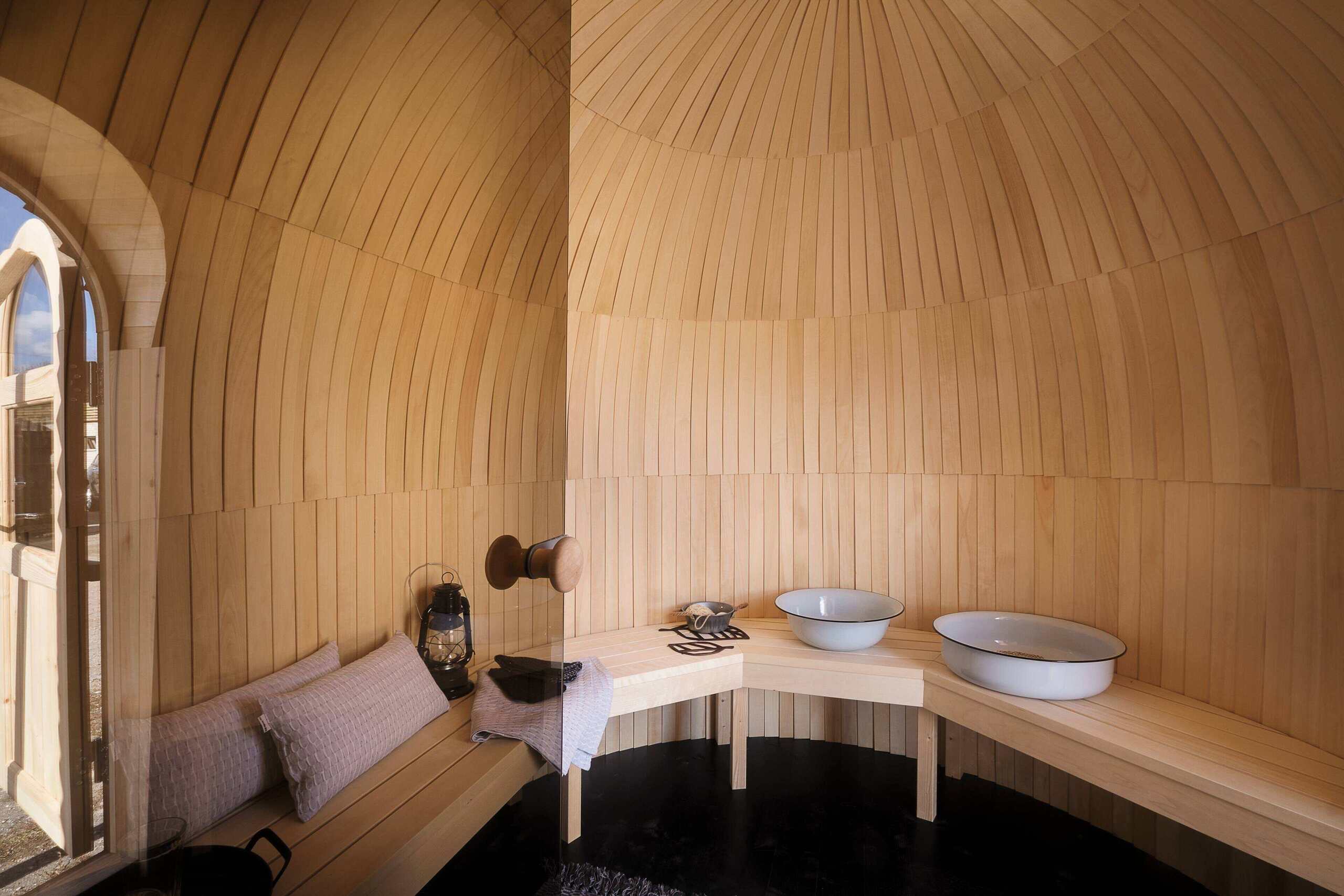 The relaxation room inside a Natural-finish Double Iglusauna — light aspen domed ceiling, aspen benches with a lantern and white enamel wash basins, the arched window glowing with daylight on the left, the glass sauna door visible on the right