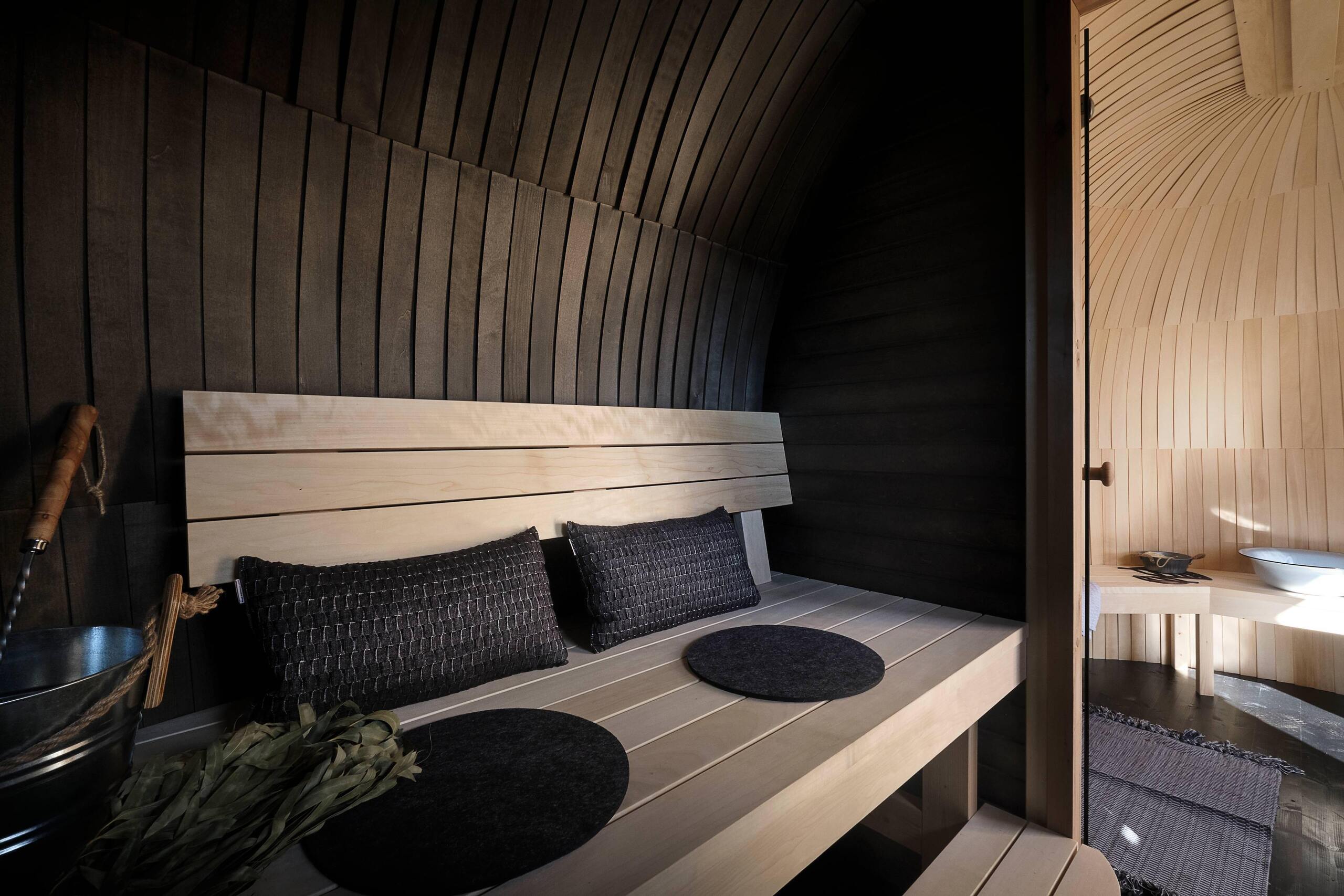 The sauna room inside a Smoke Sauna Black Double Iglusauna — darkened aspen walls and domed ceiling, lighter aspen benches with cushions, a sauna whisk and water bucket nearby, the lighter relaxation room visible through the glass door on the right