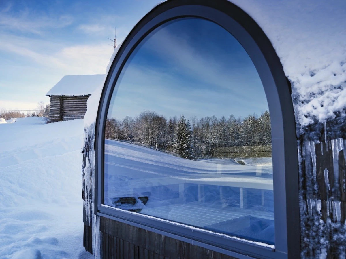 The arched panoramic window of an Ebony Iglusauna reflecting a snow-covered forest, with frost clinging to the shingles and icicles forming along the roofline