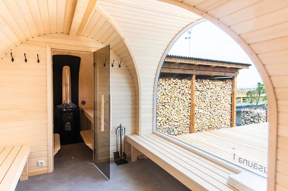 The relaxation room of a Natural-finish Panoramic Iglusauna — light aspen walls, aspen benches with folded white towels, the arched ceiling sweeping overhead, and a wide window looking out toward a marina