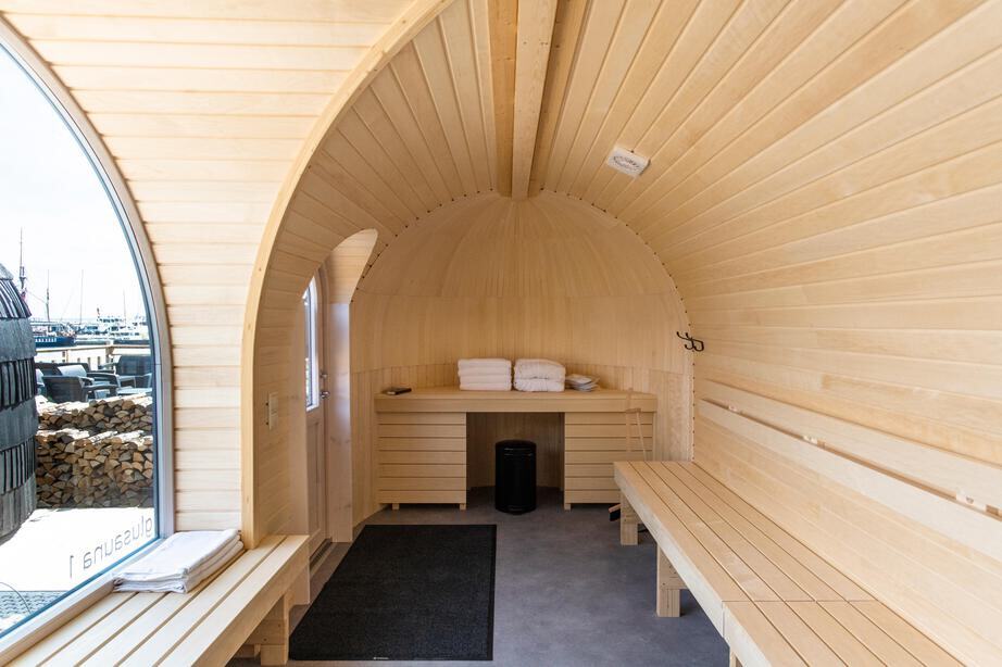Inside the sauna room of an Iglusauna — pale aspen walls and benches, a glass-doored sauna with a wood-burning stove visible, and the panoramic window looking out toward a neatly stacked wood store