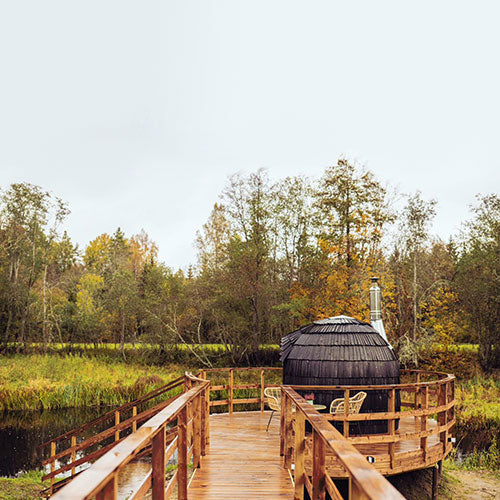 An Ebony Single Iglusauna on a wooden boardwalk extending over wetlands, surrounded by autumn trees turning gold