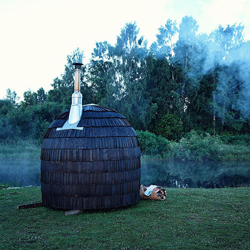 An Ebony Single Iglusauna by a lake at dusk, smoke rising from the wood-fired chimney, a small pile of firewood resting at the base, soft blue twilight settling over the water
