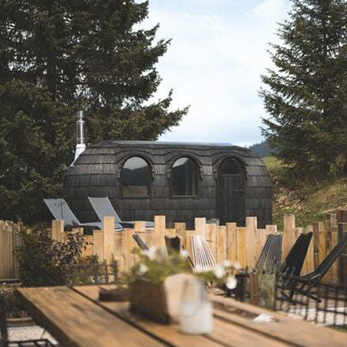 An Ebony Triple Iglusauna at a mountain resort setting, wooden fence and sun loungers in the foreground, evergreen trees behind, the elongated rounded form set against the landscape