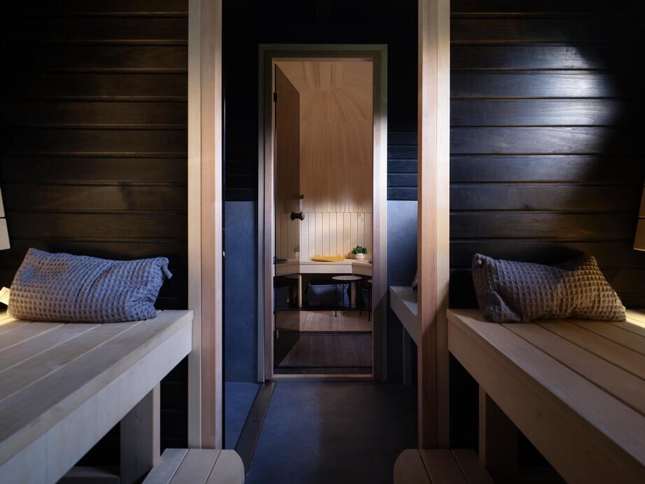 Looking from the Smoke Sauna Black sauna room through an open glass door into the lighter washing room beyond — dark stained aspen walls in the foreground, light aspen benches with cushions, the warmer light of the next room drawing you forward