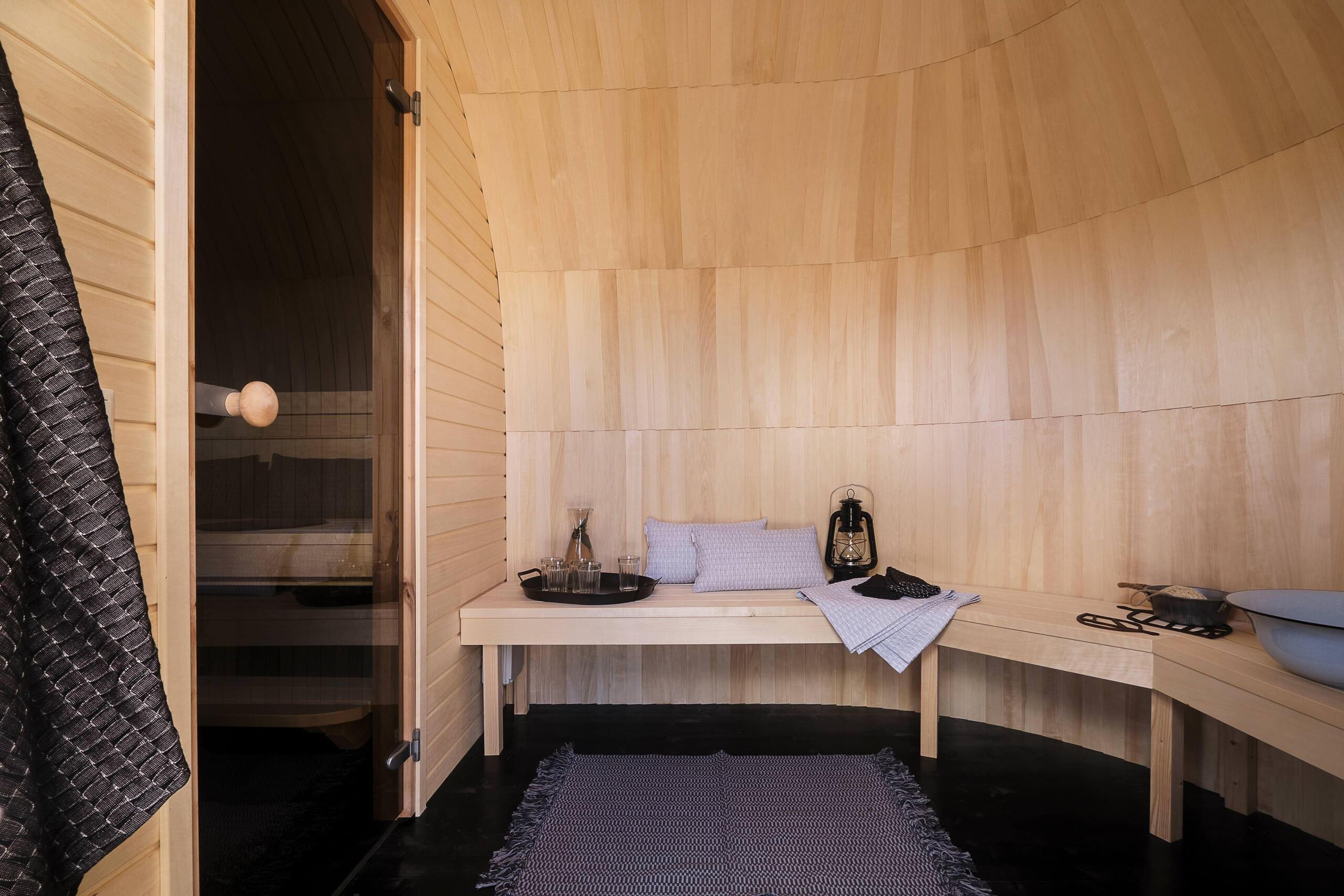 The changing room inside a Natural-finish Triple Iglusauna — light aspen walls and arched ceiling, a wooden bench with cushions and a lantern, glasses of water on a tray, a glass sauna door visible on the left leading to the next room