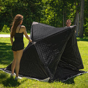 Two people pulling the tent open on grass, the structure about half erected