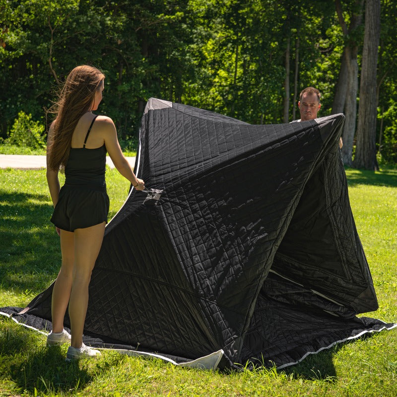 Two people pulling the tent open on grass, the structure about half erected