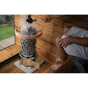 Two people inside a tent sauna, one of them pouring water over the heater's stone column